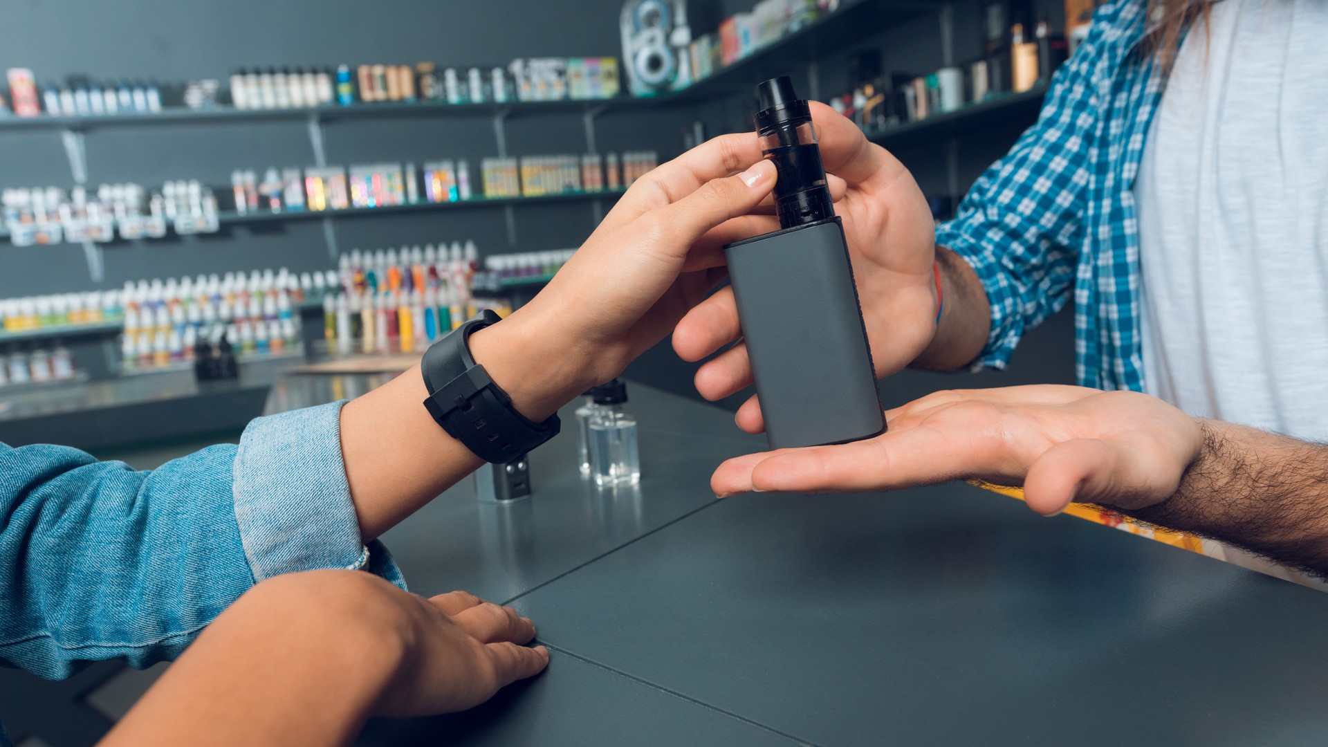 The girl came to the vapeshop. She talks with the seller - a tall man with long hair and a beard. The store has a large selection of electronic cigarettes.