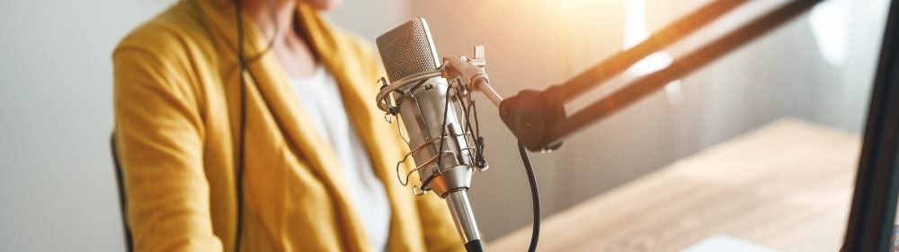 Una toma del pecho y el torso de un podcaster con una chaqueta amarilla y una camisa blanca frente a un micrófono en una historia sobre los esfuerzos de descubrimiento de podcasts de YouTube.