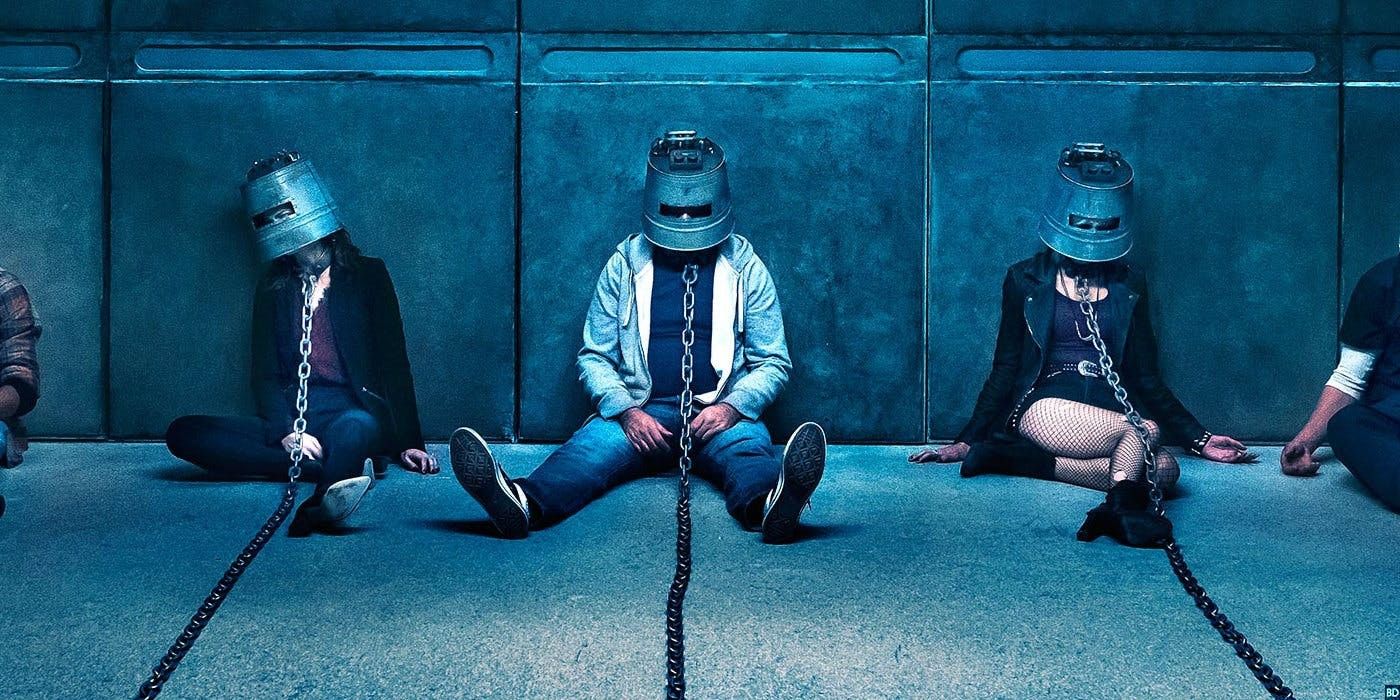 La gente se sienta en el suelo con cascos de metal encadenados a algo fuera de la pantalla en 'Jigsaw'.