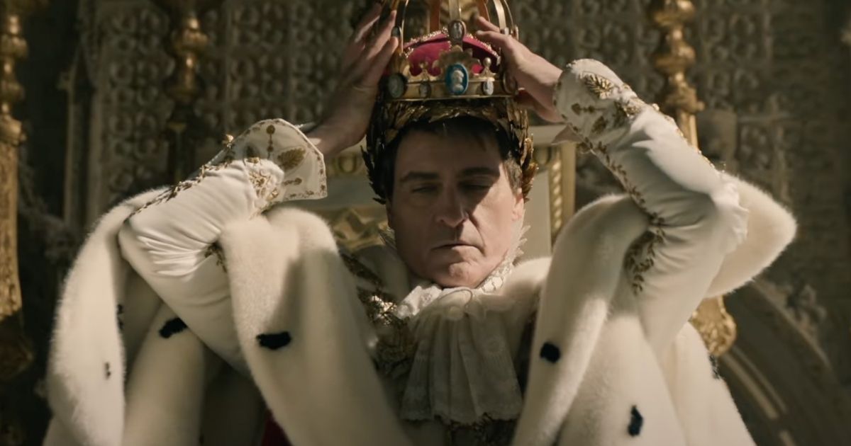 Joaquin Phoenix como Napoleón, poniéndose su corona mientras está sentado en un trono vistiendo un lujoso traje blanco con piezas de oro adheridas.