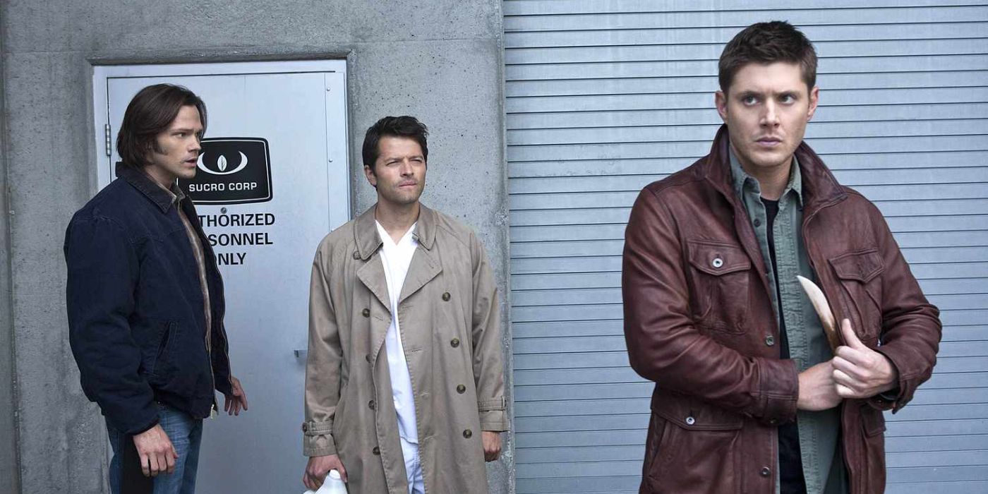 Dean Winchester pickets a knife while Sam and Cas stand behind him in front of a building's restricted entry.
